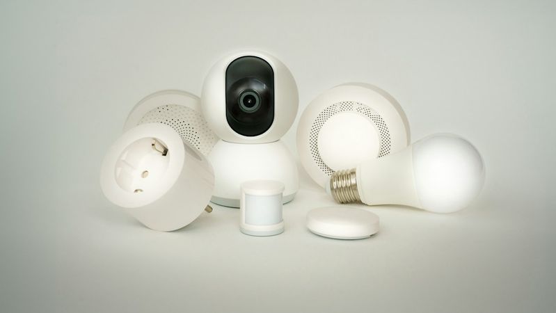 The image features a collection of smart home devices, including a security camera, smart light bulb, motion sensor, and smart plug, all designed in a sleek white aesthetic. These devices are arranged artistically on a neutral background, highlighting their modern technology.