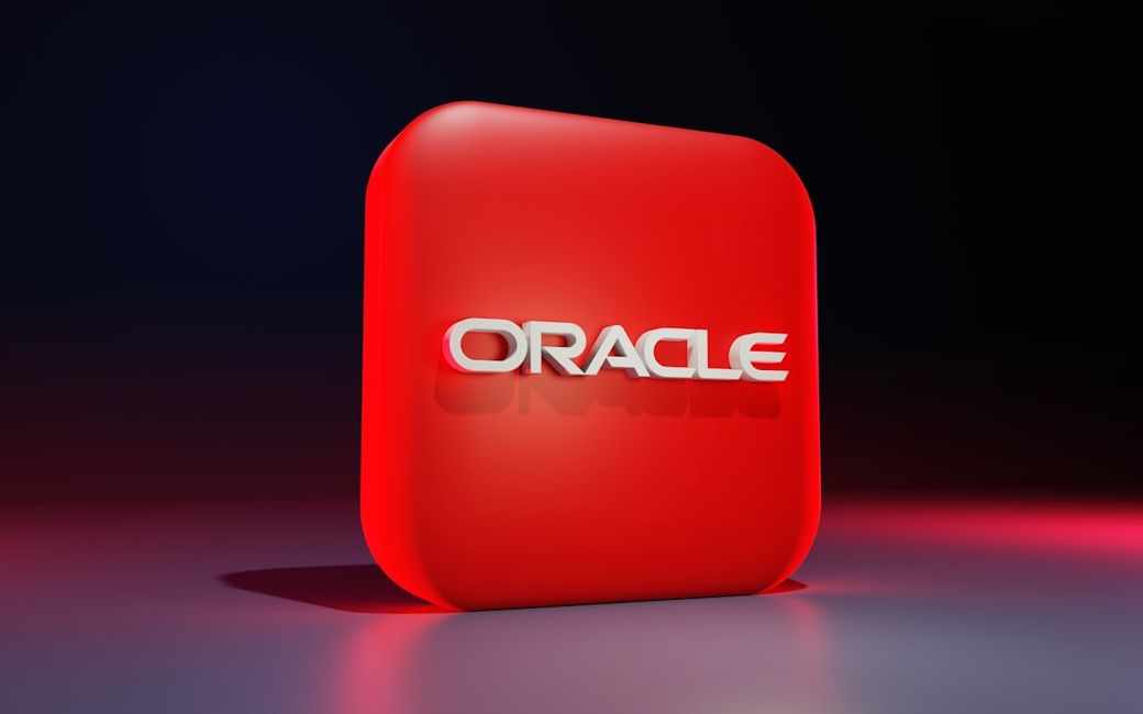 The image features a prominently displayed three-dimensional logo of Oracle, set against a gradient background. The logo is rendered in white text on a vibrant red square platform. — Credit: Photo by BoliviaInteligente on Unsplash