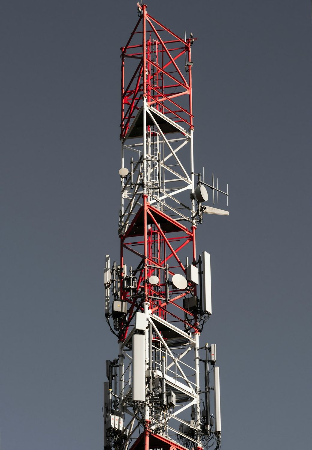The image showcases a telecommunications tower standing against a clear sky. The structure features a combination of red and white colors, equipped with various antennas and communication devices. — Credit: Photo by Rayyân on Unsplash
