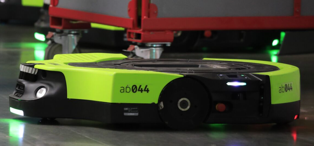 The image shows a robotic device on a smooth floor in a warehouse or industrial setting. The robot is designed for automated tasks, likely for transporting goods or materials.