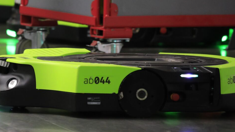 The image shows a robotic device on a smooth floor in a warehouse or industrial setting. The robot is designed for automated tasks, likely for transporting goods or materials.