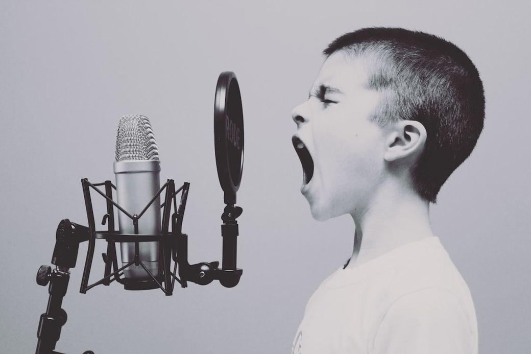 A young boy is passionately singing or yelling into a microphone, showcasing raw emotion and enthusiasm. The image is monochromatic, emphasizing the boy's expression and the microphone setup. — Credit: Photo by Jason Rosewell on Unsplash