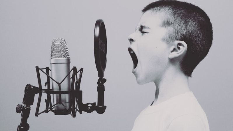 A young boy is passionately singing or yelling into a microphone, showcasing raw emotion and enthusiasm. The image is monochromatic, emphasizing the boy's expression and the microphone setup.