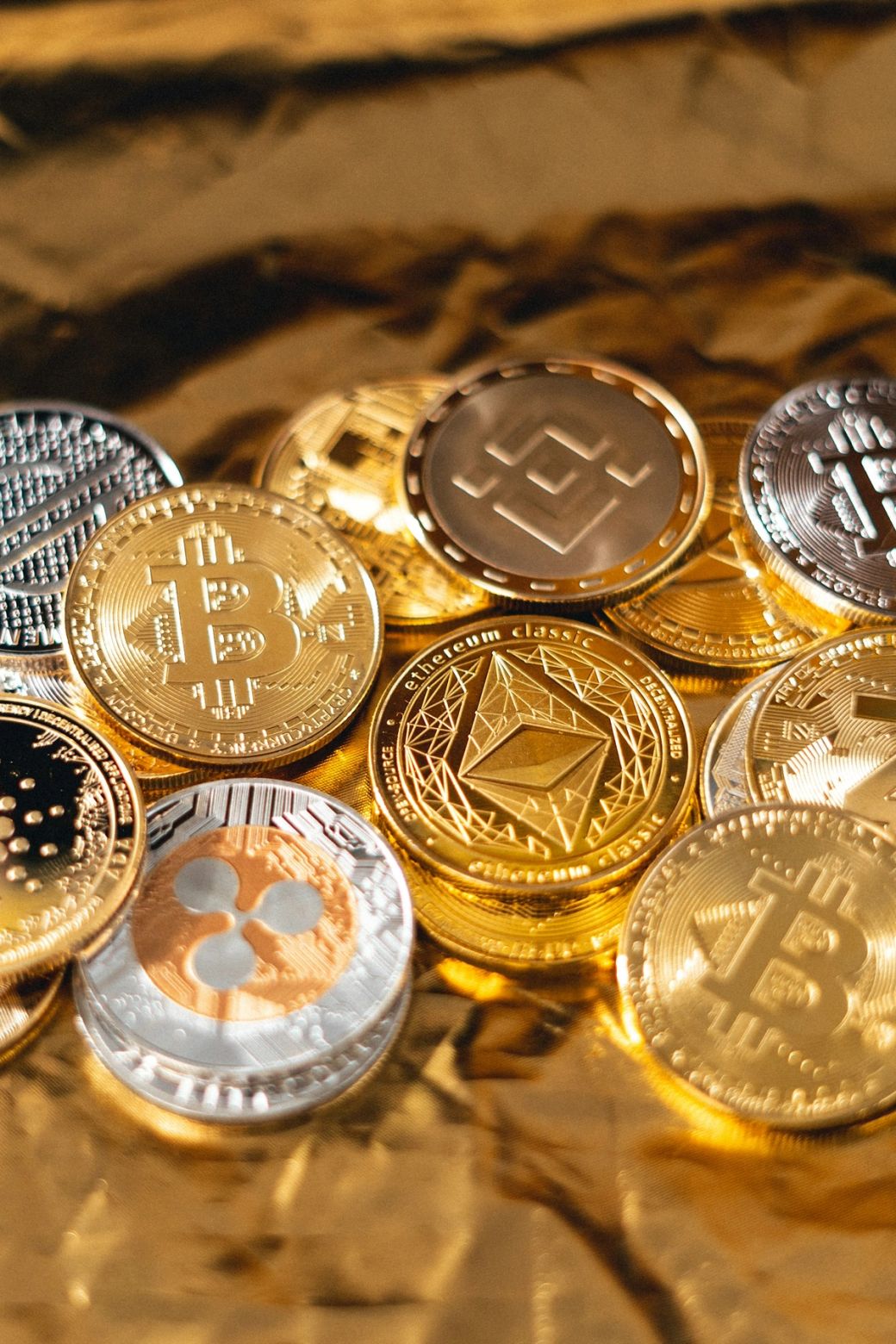 The image features a close-up view of various cryptocurrency coins, prominently displaying Bitcoin, Ethereum Classic, and Binance logos. The coins are stacked together, showcasing their detailed designs against a slightly blurred background. — Credit: Photo by Art Rachen / Unsplash
