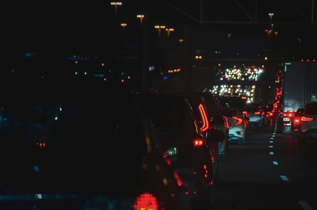The image shows a congested highway at night, with a line of cars illuminated by their tail lights. The background is a blur of colorful traffic lights, indicating a heavy flow of vehicles. — Credit: Photo by Hooman R. / Unsplash