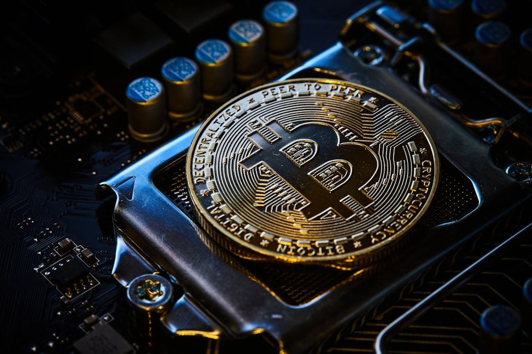 The image displays a close-up view of a Bitcoin token positioned on a computer circuit board. The shiny gold surface of the token prominently features the Bitcoin logo alongside intricate details highlighting its unique design. — Credit: Photo by Michael Förtsch on Unsplash