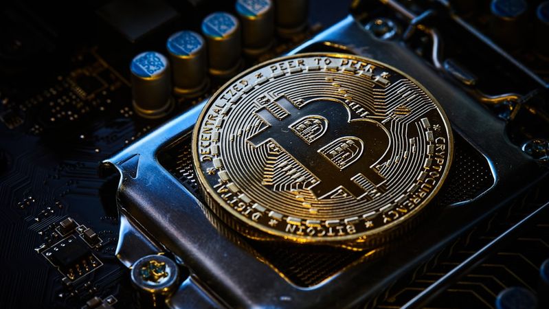 The image displays a close-up view of a Bitcoin token positioned on a computer circuit board. The shiny gold surface of the token prominently features the Bitcoin logo alongside intricate details highlighting its unique design.