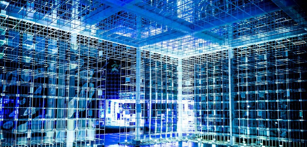 The image depicts an abstract architectural space comprised of interconnected glass-like structures illuminated by blue lighting. The environment conveys a futuristic and high-tech aesthetic. — Credit: Photo by Kelvin Ang on Unsplash