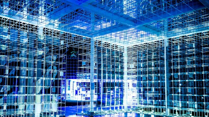 The image depicts an abstract architectural space comprised of interconnected glass-like structures illuminated by blue lighting. The environment conveys a futuristic and high-tech aesthetic.