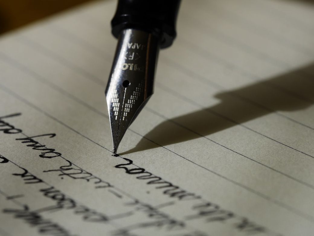 The image features a close-up view of a fountain pen poised above a sheet of lined paper with handwritten text. The focus is on the pen's nib, highlighting the elegance of writing. — Credit: Photo by Aaron Burden on Unsplash