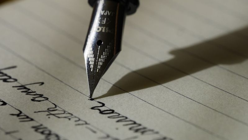 The image features a close-up view of a fountain pen poised above a sheet of lined paper with handwritten text. The focus is on the pen's nib, highlighting the elegance of writing.