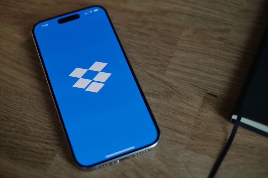 The image shows a smartphone displaying the Dropbox logo on a blue screen, placed on a wooden surface next to a notebook. The clean and minimalist design highlights the app's branding. — Credit: Photo by appshunter.io on Unsplash