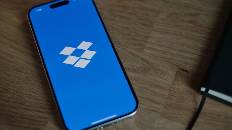 The image shows a smartphone displaying the Dropbox logo on a blue screen, placed on a wooden surface next to a notebook. The clean and minimalist design highlights the app's branding.