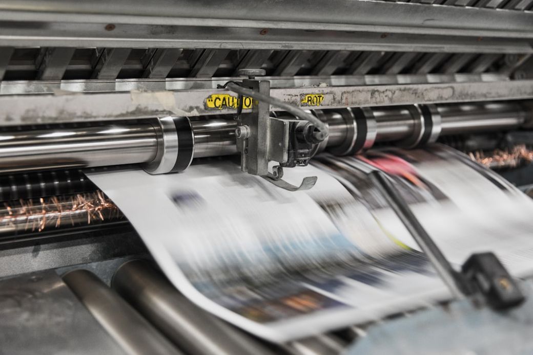 The image shows a printing press in operation, with sheets of printed material being fed through the machine. The motion blur indicates the speed at which the printing process occurs. — Credit: Photo by Bank Phrom on Unsplash