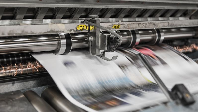 The image shows a printing press in operation, with sheets of printed material being fed through the machine. The motion blur indicates the speed at which the printing process occurs.