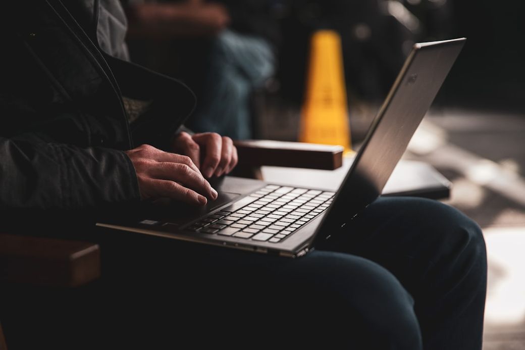 An individual sits on a wooden chair typing on a laptop in a casual setting. The focus is on their hands and the keyboard, suggesting a moment of productivity or concentration. — Credit: Photo by LOGAN WEAVER | @LGNWVR on Unsplash