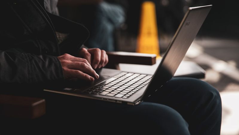 An individual sits on a wooden chair typing on a laptop in a casual setting. The focus is on their hands and the keyboard, suggesting a moment of productivity or concentration.