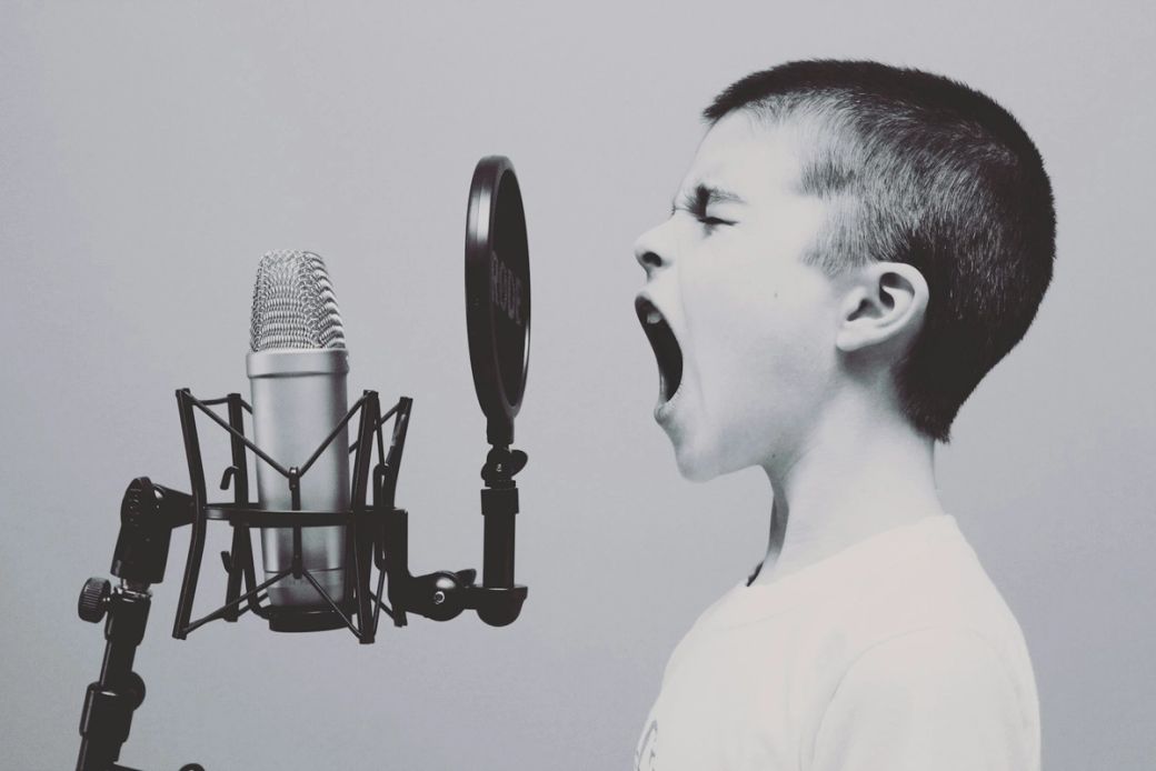 A young boy passionately singing or shouting into a microphone, capturing a moment of expression in a studio setting. The image features a close-up of the boy's face alongside a professional microphone. — Credit: Photo by Jason Rosewell / Unsplash