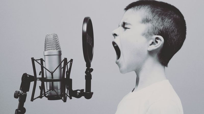 A young boy passionately singing or shouting into a microphone, capturing a moment of expression in a studio setting. The image features a close-up of the boy's face alongside a professional microphone.