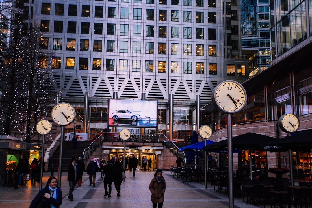 The image depicts a bustling urban plaza featuring several prominent clocks and a large advertising screen displaying a car. People are walking amidst the modern architecture and vibrant atmosphere of the area.
