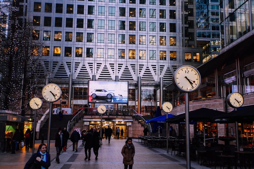 The image depicts a bustling urban plaza featuring several prominent clocks and a large advertising screen displaying a car. People are walking amidst the modern architecture and vibrant atmosphere of the area. — Credit: Photo by Laureen Missaire on Unsplash