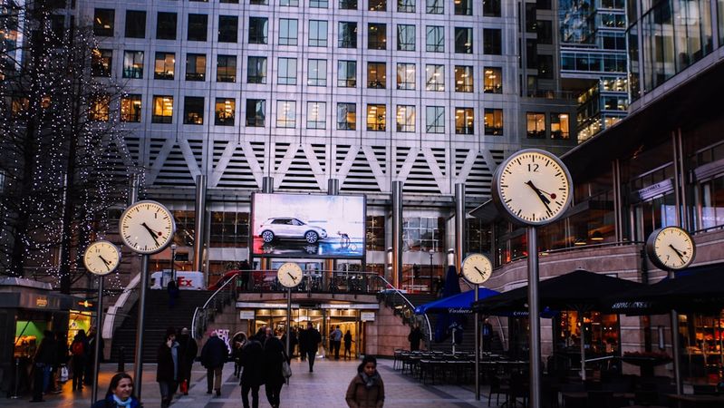 The image depicts a bustling urban plaza featuring several prominent clocks and a large advertising screen displaying a car. People are walking amidst the modern architecture and vibrant atmosphere of the area.