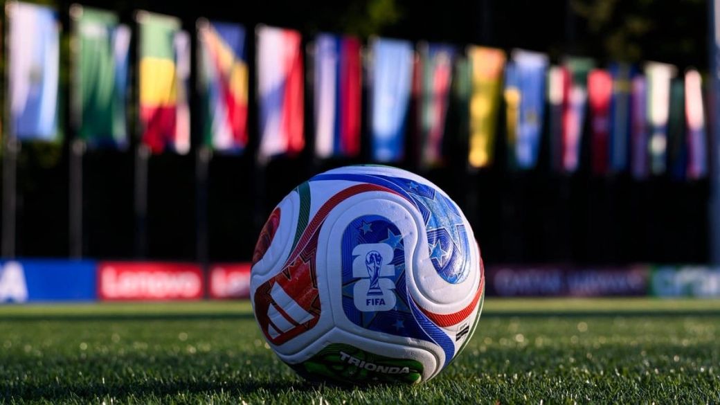 A close-up view of a FIFA-branded soccer ball situated on a grassy field, with a backdrop of flags from various countries. The scene captures the essence of international sports competition.