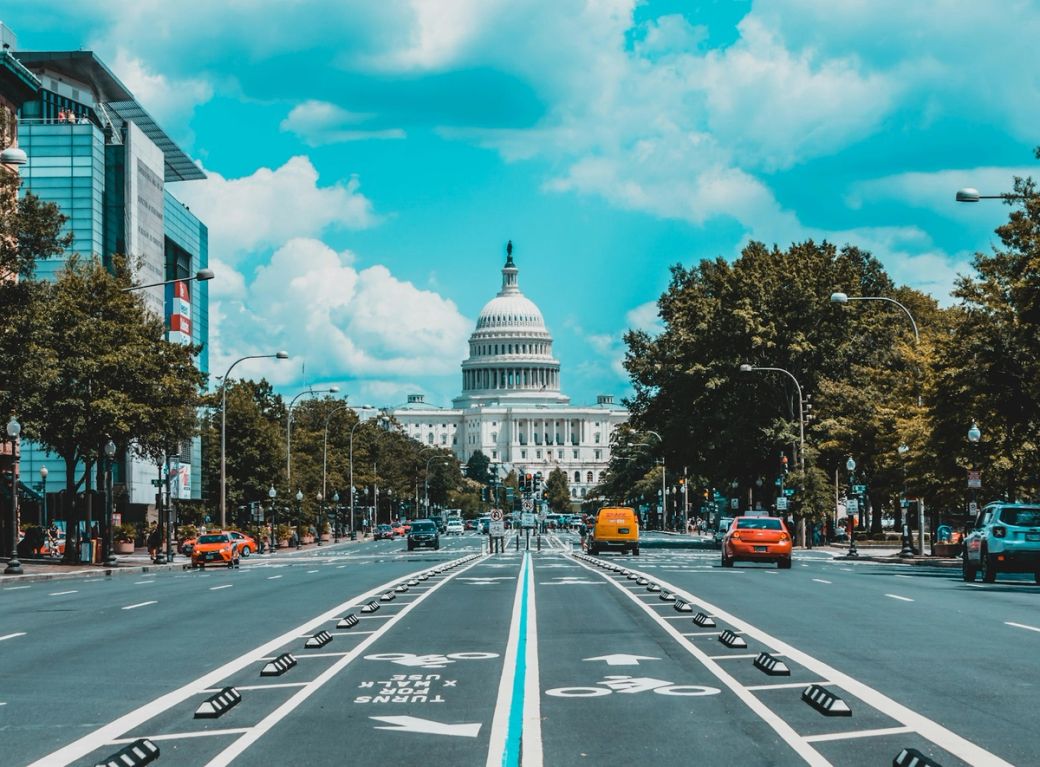 The image depicts a wide street in Washington, D.C., leading toward the United States Capitol building. Vehicles are seen on the road, and the sky features vibrant clouds, contributing to the overall bright atmosphere of the scene. — Credit: Photo by Jorge Alcala / Unsplash