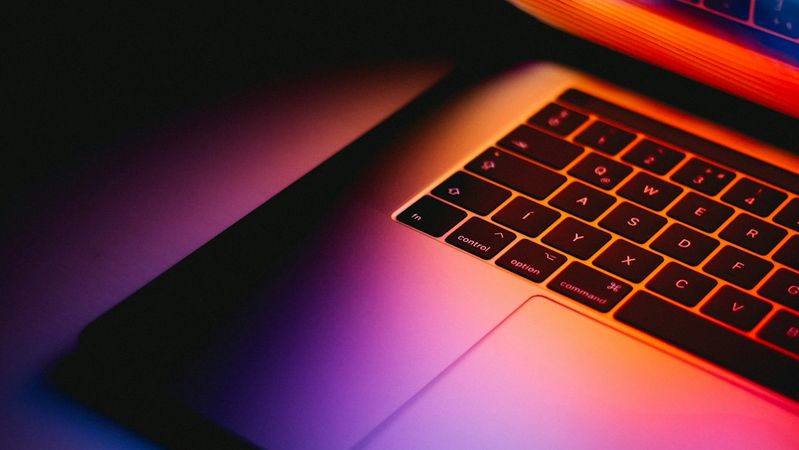 The image features a close-up view of a laptop with a colorful display, the light reflecting on its keyboard and surface. The scene is set against a dark background, creating a dramatic contrast with the vibrant colors emitted from the screen.