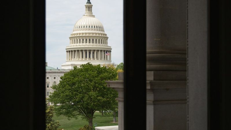 The image captures the United States Capitol building as seen through a doorway. The view highlights the iconic dome and surrounding greenery, framing the structure amidst a partially shaded setting.
