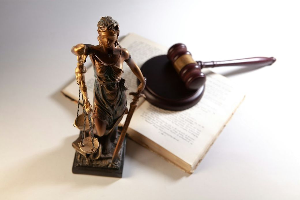 The image depicts a figurine of Lady Justice, blindfolded and holding scales, alongside a gavel and an open law book. The arrangement symbolizes the legal profession and the concept of justice. — Credit: Photo by Sasun Bughdaryan on Unsplash