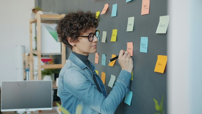 A young woman with curly hair and glasses is writing on colorful sticky notes attached to a black wall in a modern office space. She is focused on her task, indicating a collaborative work environment.