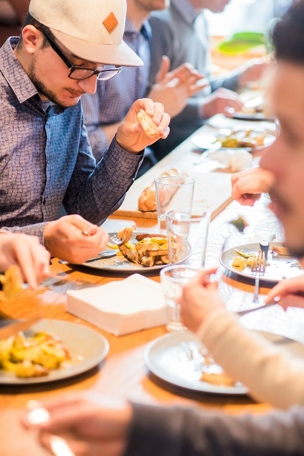 The image depicts a group of people gathered around a wooden table enjoying a meal. One individual is seen focusing on their plate while others are engaged in conversation and eating. — Credit: Photo by Proxyclick Visitor Management System on Unsplash