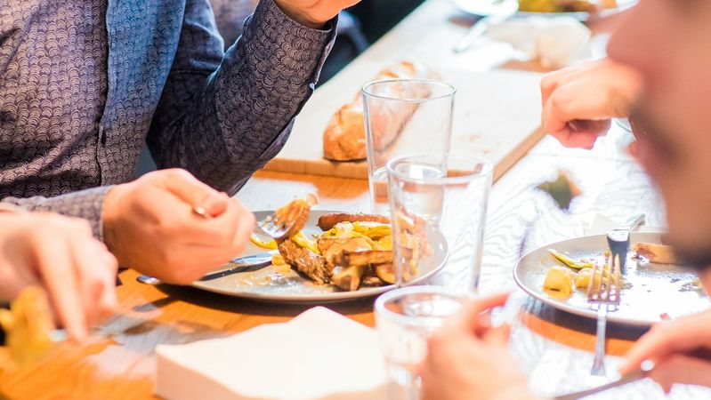 The image depicts a group of people gathered around a wooden table enjoying a meal. One individual is seen focusing on their plate while others are engaged in conversation and eating.