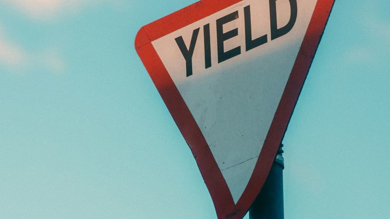 A close-up image of a yield traffic sign against a clear blue sky. The sign is prominently displayed, featuring a red triangle shape with the word 'YIELD' in bold black letters.