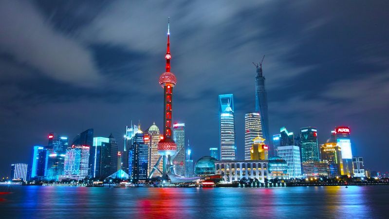 The image showcases the skyline of Shanghai at night, featuring illuminated skyscrapers and the iconic Oriental Pearl Tower. The vibrant cityscape reflects in the water, highlighting the modern architecture and urban landscape of one of China's major financial hubs.
