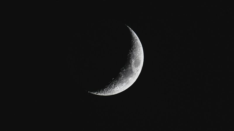 A close-up view of a crescent moon against a dark background, showcasing its illuminated and shadowed features. The image captures the delicate curvature and surface details of the lunar surface.