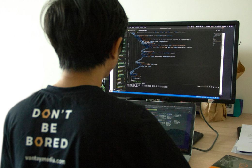 A person is seen programming at a desk with two monitors. One monitor displays lines of code, while the other shows a focused view of the coding activity, suggesting a software development environment. — Credit: Photo by Van Tay Media / Unsplash