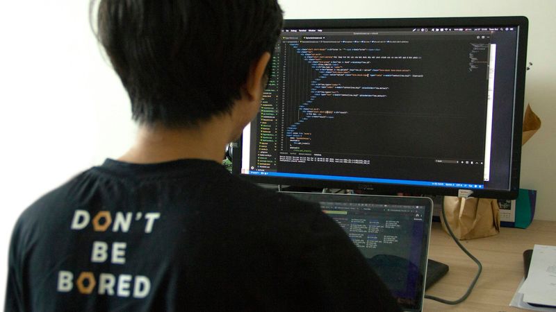A person is seen programming at a desk with two monitors. One monitor displays lines of code, while the other shows a focused view of the coding activity, suggesting a software development environment.