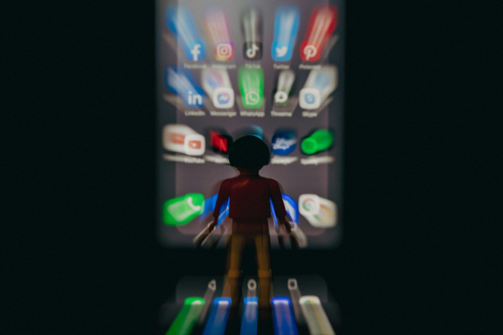 The image features a blurred figure of a toy character standing in front of a smartphone screen displaying various social media app icons. The focus is on the silhouette of the character, emphasizing the contrast between the real-world object and the digital interface.