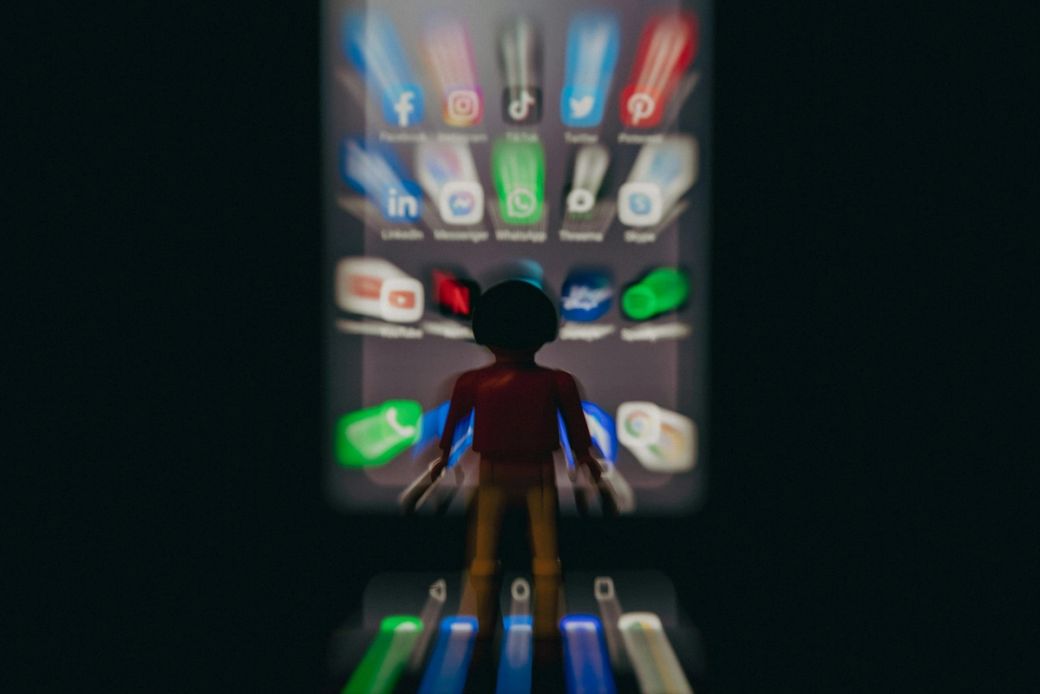 The image features a blurred figure of a toy character standing in front of a smartphone screen displaying various social media app icons. The focus is on the silhouette of the character, emphasizing the contrast between the real-world object and the digital interface. — Credit: Photo by Florian Schmetz / Unsplash