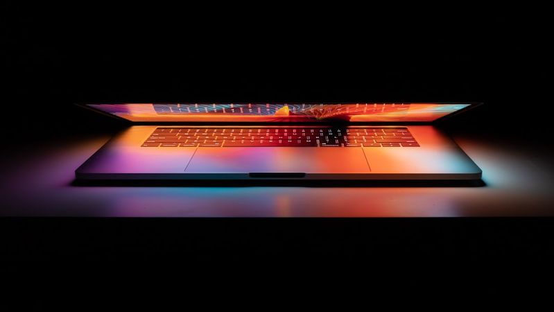 The image features a laptop with an open screen, showcasing a colorful display against a dark background. The lighting creates a striking contrast with vibrant hues illuminating the laptop's body.