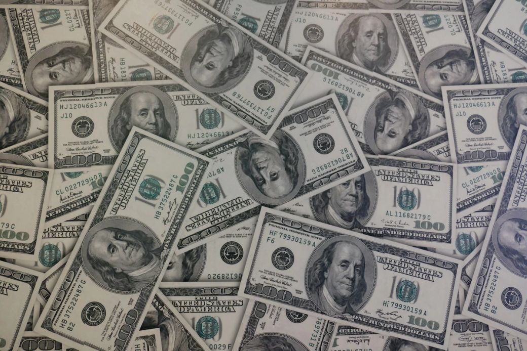 A close-up view of numerous hundred-dollar bills scattered across a surface. The image highlights the intricate details of the currency, showcasing the iconic portrait and design elements. — Credit: Photo by Mackenzie Marco / Unsplash