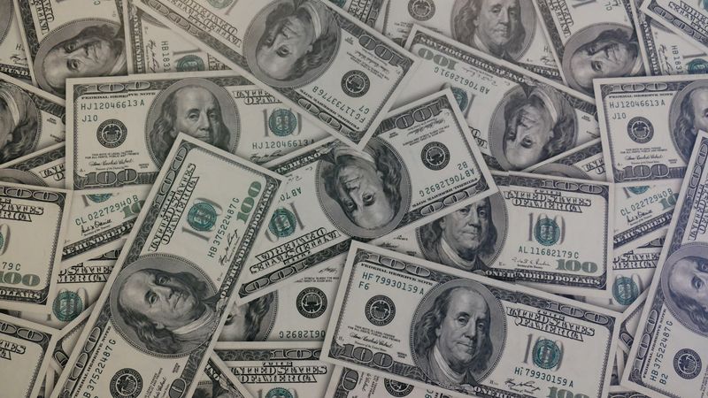 A close-up view of numerous hundred-dollar bills scattered across a surface. The image highlights the intricate details of the currency, showcasing the iconic portrait and design elements.