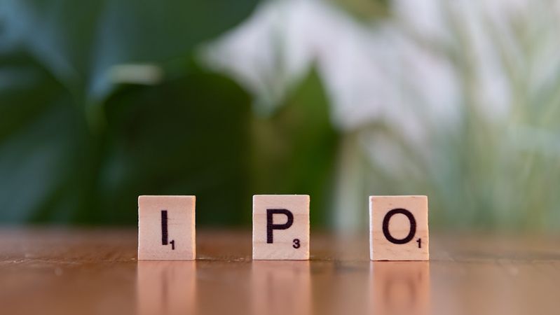 The image shows wooden letter tiles spelling out 'IPO' against a blurred green background. The tiles are positioned on a wooden surface, suggesting a focus on finance or investment themes.