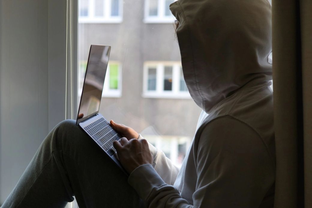 A person with a hooded sweatshirt sits by a window, working on a laptop. The background shows a blurred cityscape with multiple windowed buildings. — Credit: Photo by Kaptured by Kasia / Unsplash