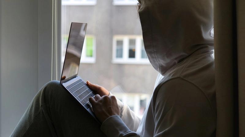 A person with a hooded sweatshirt sits by a window, working on a laptop. The background shows a blurred cityscape with multiple windowed buildings.