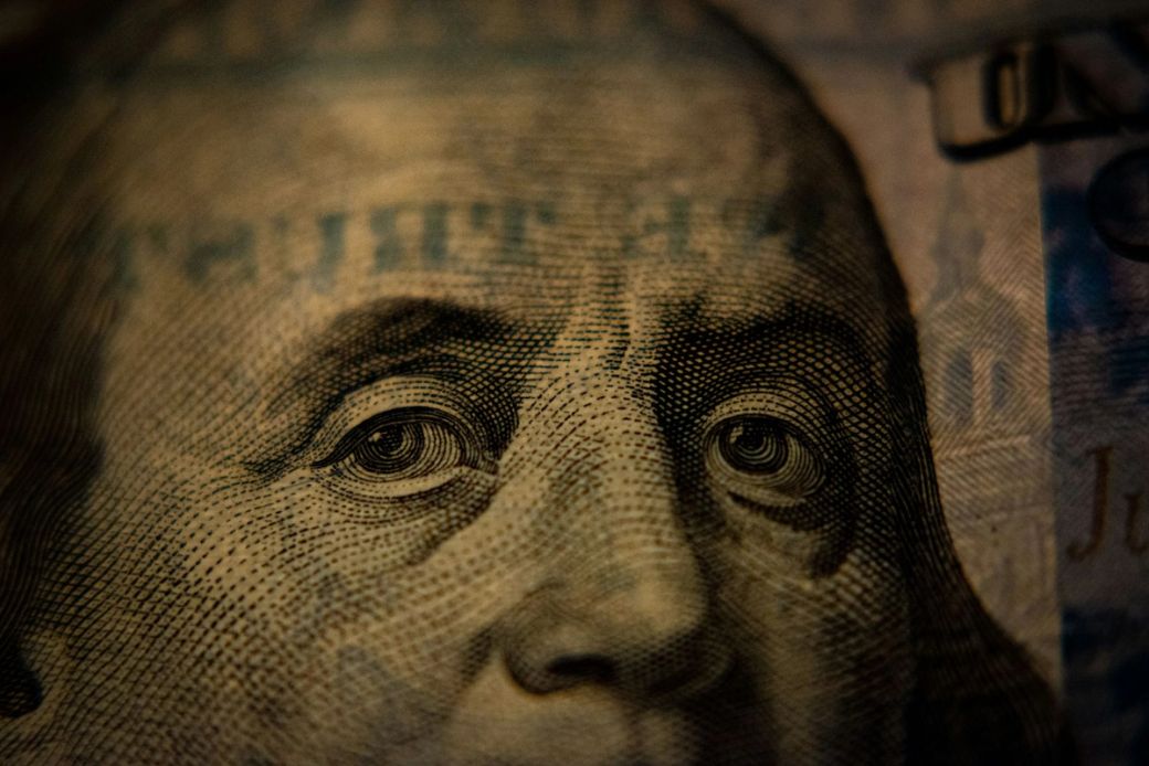The image features a close-up of a Benjamin Franklin portrait printed on a US one-hundred-dollar bill, highlighting intricate details of the facial features and engraving. The lighting creates a dramatic effect on the texture of the bill. — Credit: Photo by Adam Nir / Unsplash