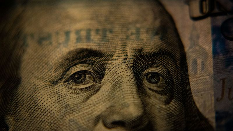 The image features a close-up of a Benjamin Franklin portrait printed on a US one-hundred-dollar bill, highlighting intricate details of the facial features and engraving. The lighting creates a dramatic effect on the texture of the bill.