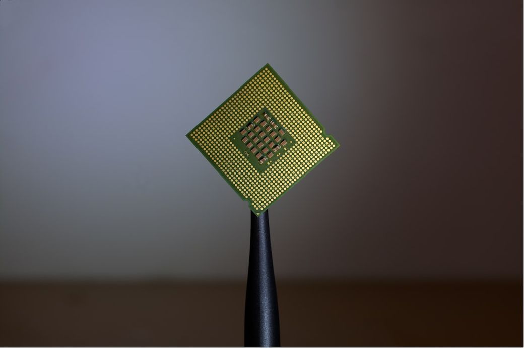 The image features a close-up of a computer processor being held by tweezers against a soft, blurred background. The processor is a small, rectangular chip with a grid of gold pins visible on its underside. — Credit: Photo by Brian Kostiuk on Unsplash